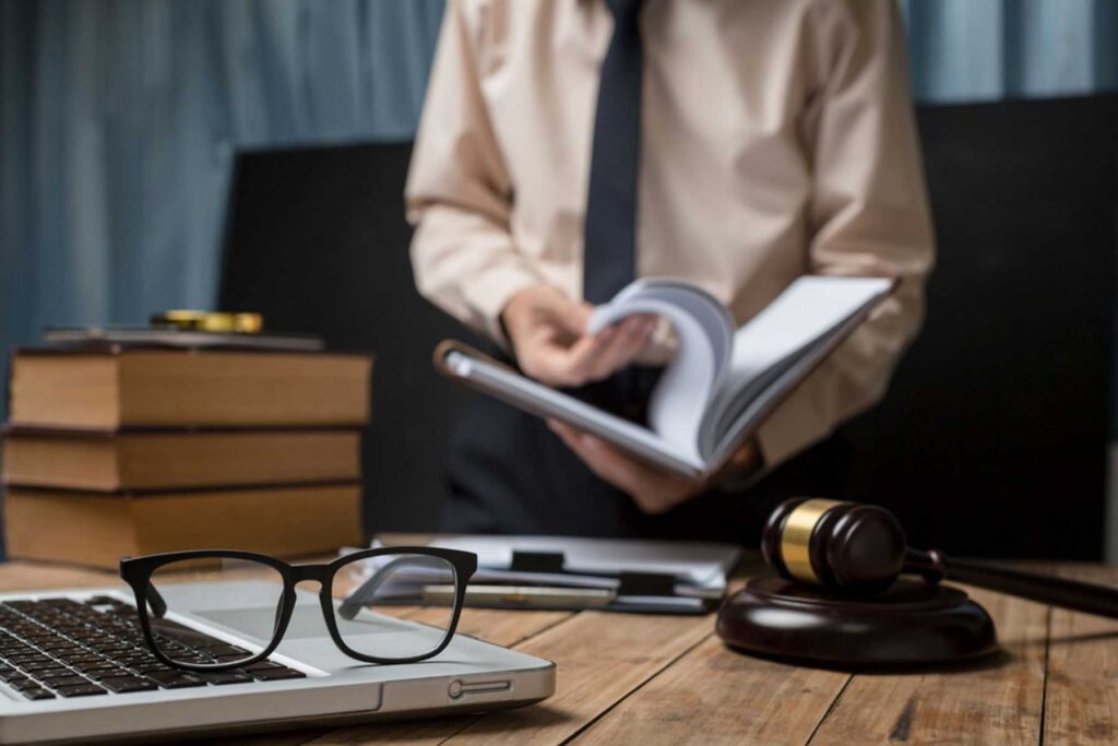 Businessman in shirt and tie reading an open book at a wooden desk with stacked books, a gavel, glasses, and a laptop